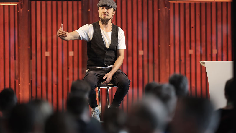 Tobias Conrad, Speaker, sitzt auf einem Hocker auf einer Bühne vor seinem Publikum.