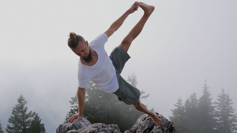 Tobias Conrad in einer Yogapose am Berg auf Felsen im Hintergrund Bäume
